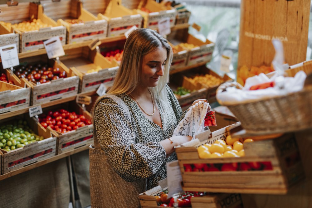 En kvinne med langt blondt hår handler ferske grønnsaker i en lokal butikk, omgitt av kasser fylt med tomater og andre fargerike grønnsaker.